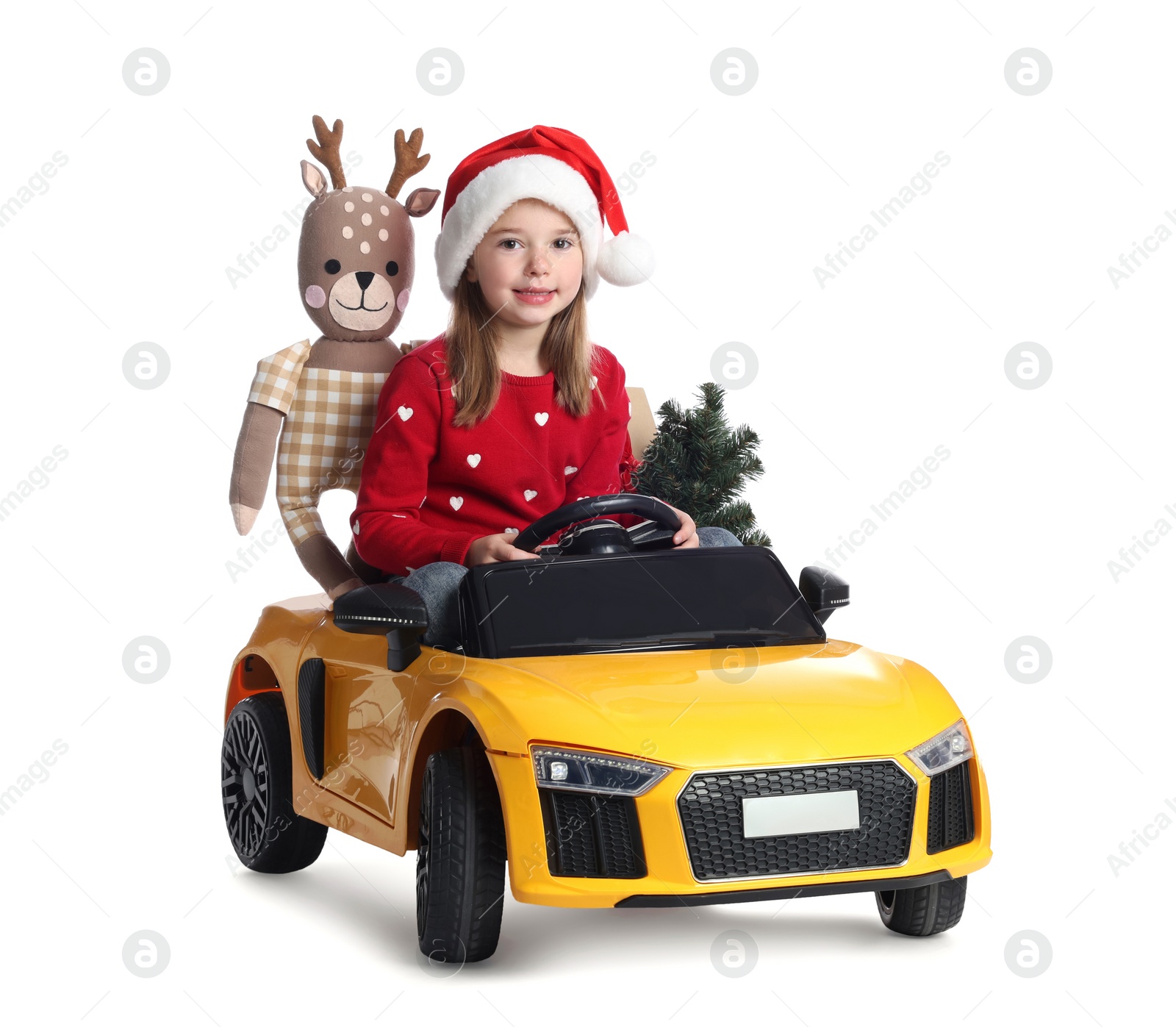 Photo of Cute little girl in Santa hat with Christmas tree and deer toy driving children's car on white background