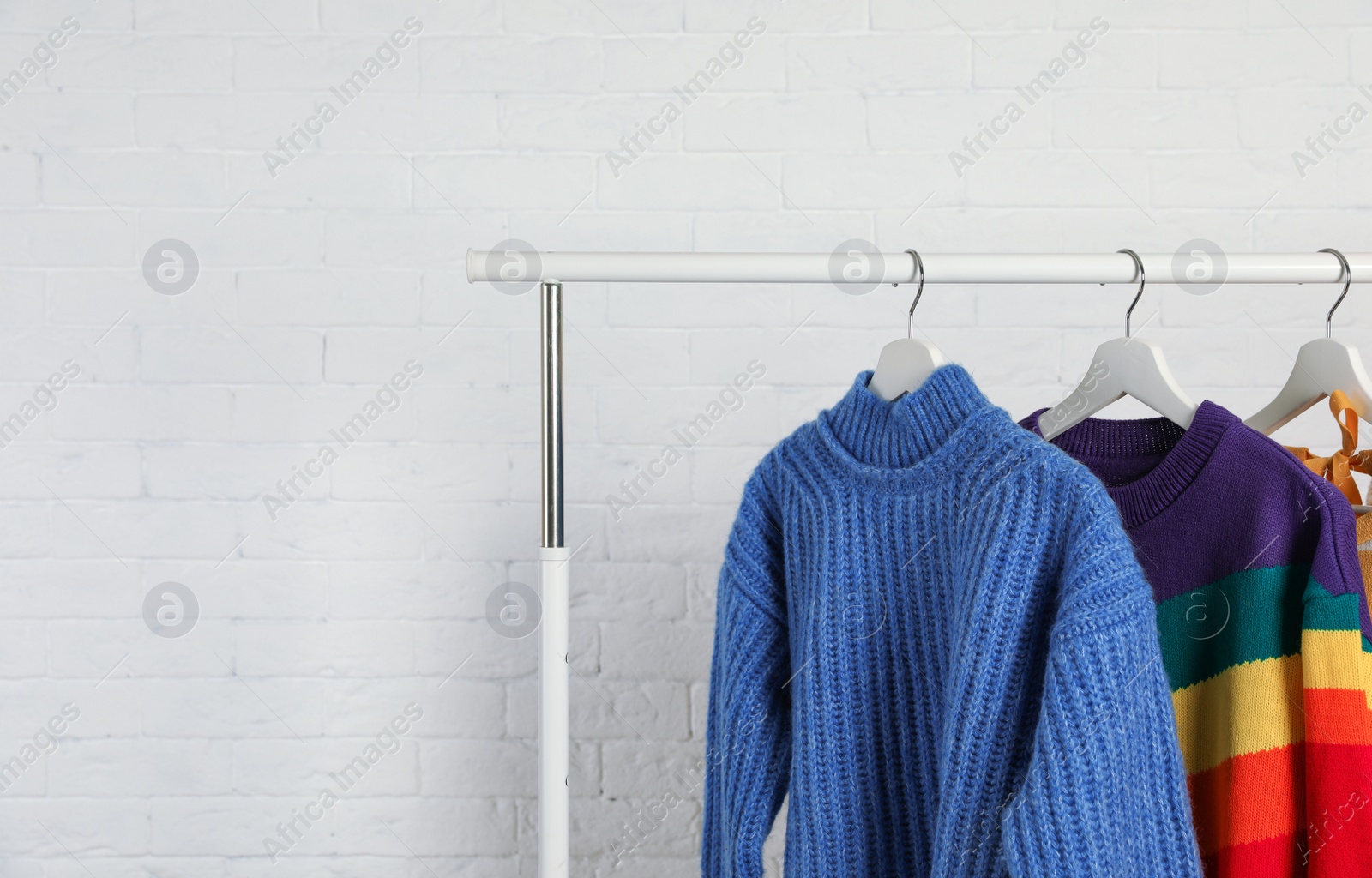 Photo of Collection of warm sweaters hanging on rack against white brick wall. Space for text