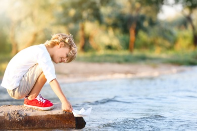 Cute little boy with paper ship at river, space for text. Child spending time in nature