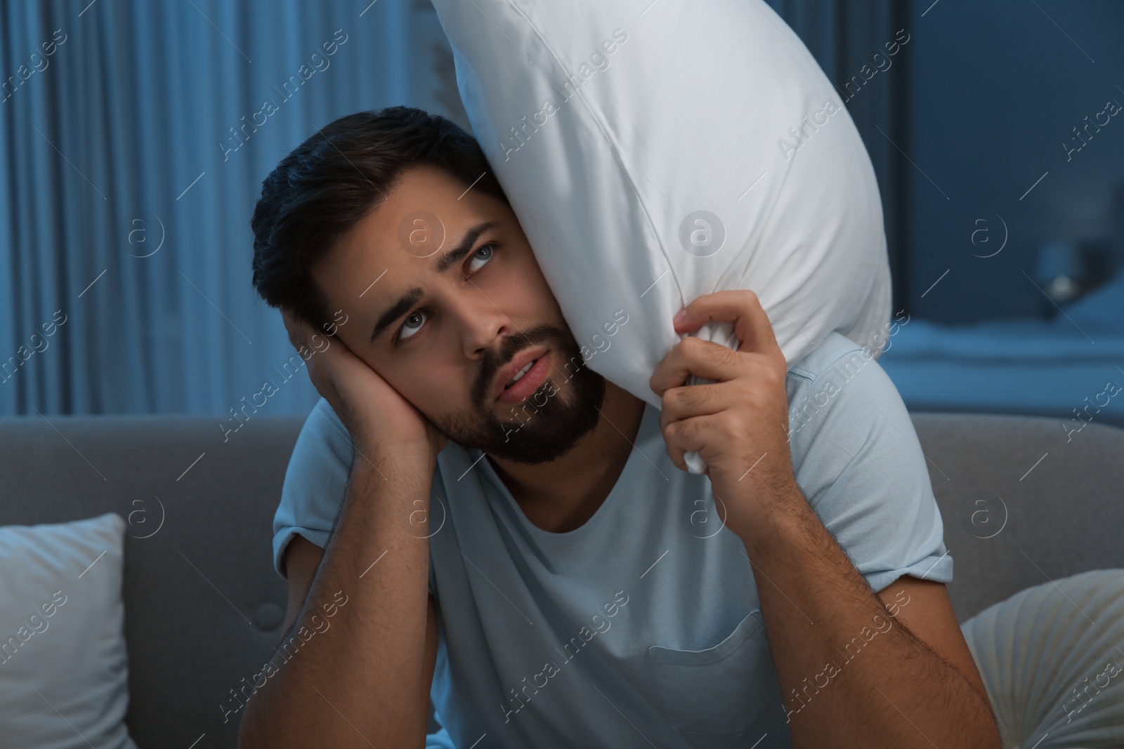 Photo of Unhappy young man covering ears in living room at night. Noisy neighbours