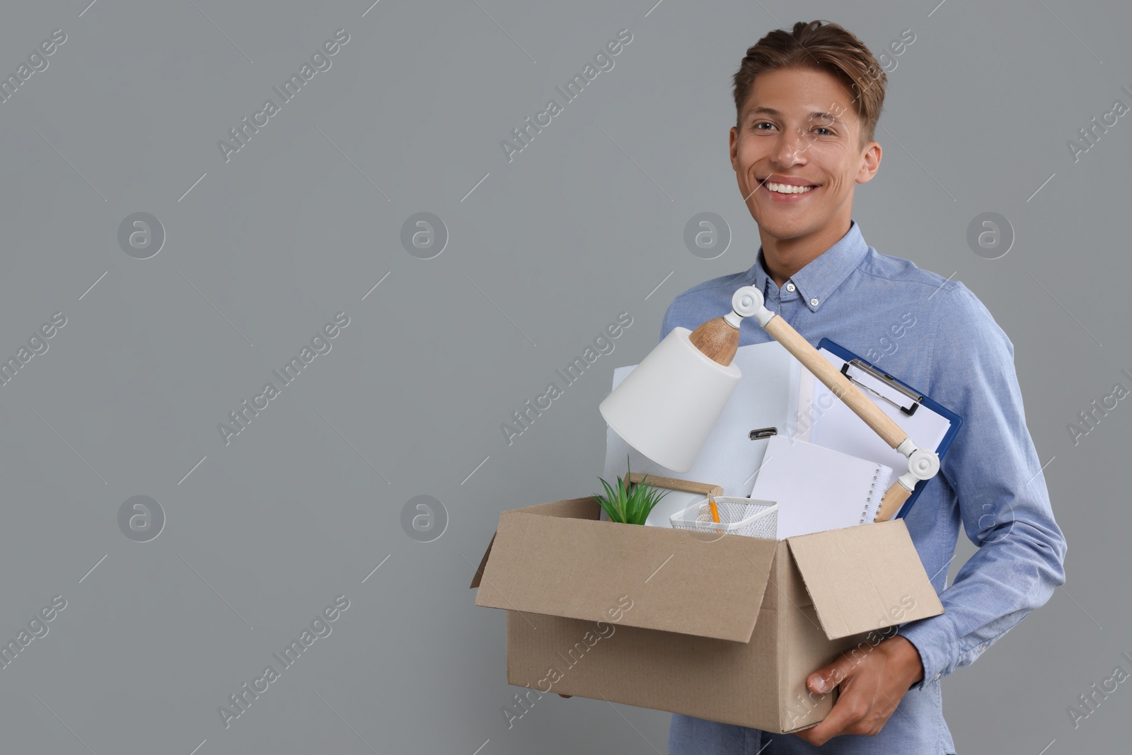 Photo of Happy unemployed young man with box of personal office belongings on grey background. Space for text