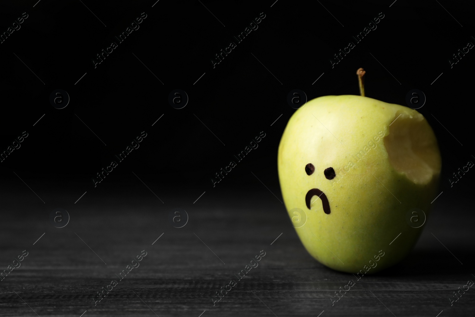 Photo of Bitten apple with drawing of unhappy face on table against dark background, space for text. Depression symptoms