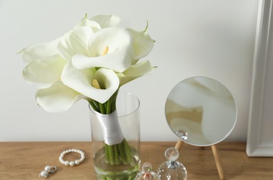 Photo of Beautiful calla lily flowers in glass vase, bottles with perfumes, jewelry and mirror on wooden table