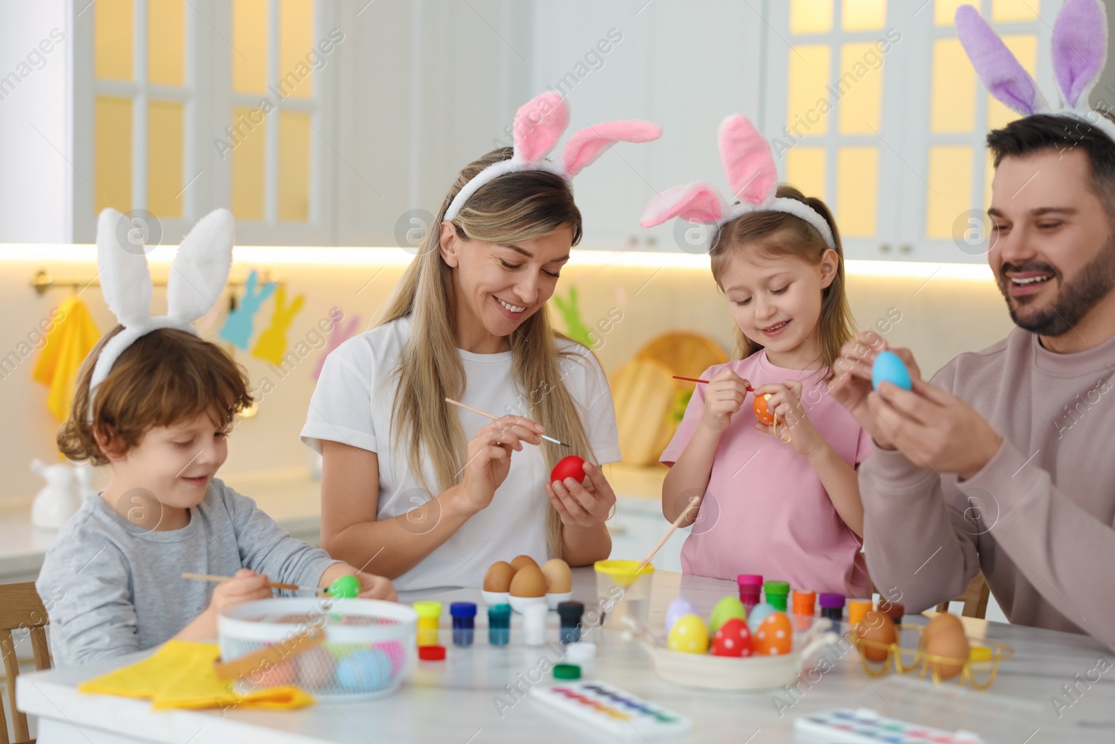 Photo of Easter celebration. Happy family with bunny ears painting eggs at white marble table in kitchen