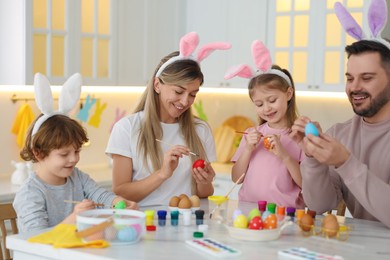 Photo of Easter celebration. Happy family with bunny ears painting eggs at white marble table in kitchen