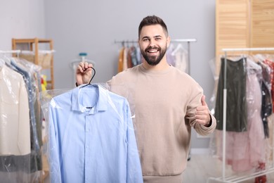 Photo of Dry-cleaning service. Happy man holding hanger with shirt in plastic bag and showing thumb up indoors