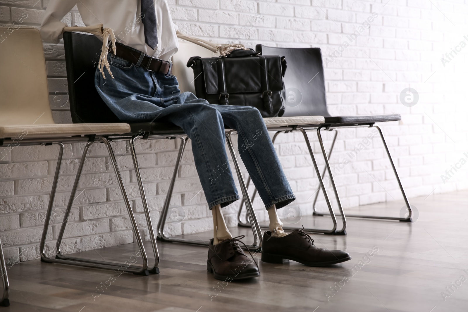 Photo of Human skeleton in office wear sitting on chair near brick wall indoors, closeup