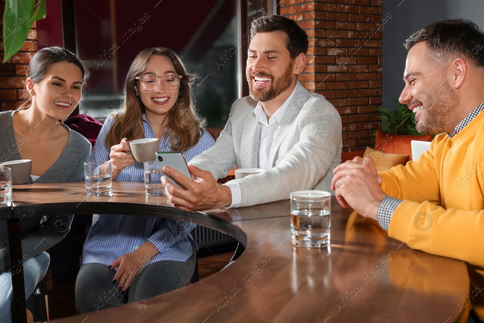 Photo of Handsome man showing something funny in smartphone to his friends in cafe