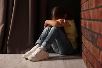 Child abuse. Upset little girl sitting on floor near brick wall indoors