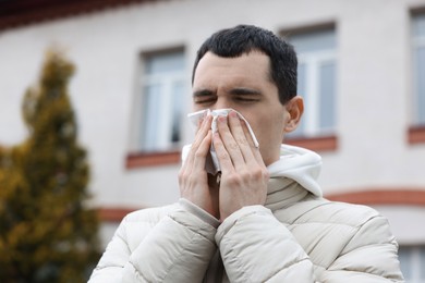 Photo of Sick young man with tissue blowing runny nose outdoors. Cold symptom