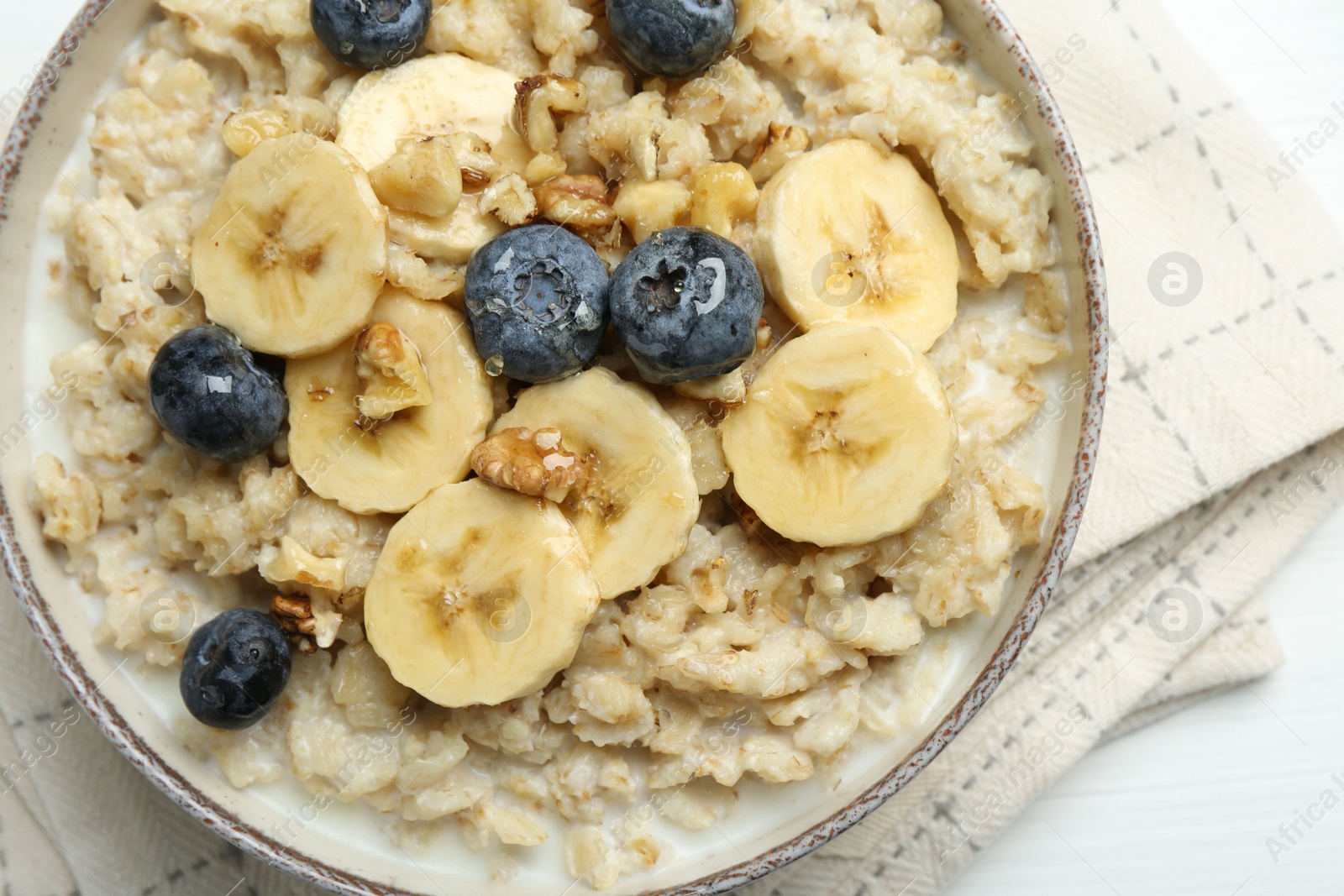 Photo of Tasty oatmeal with banana, blueberries, walnuts and honey served in bowl on white wooden table, top view