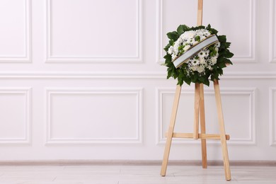 Photo of Funeral wreath of flowers with ribbon on wooden stand near white wall indoors. Space for text