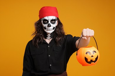 Photo of Man in scary pirate costume with skull makeup and pumpkin bucket on orange background. Halloween celebration