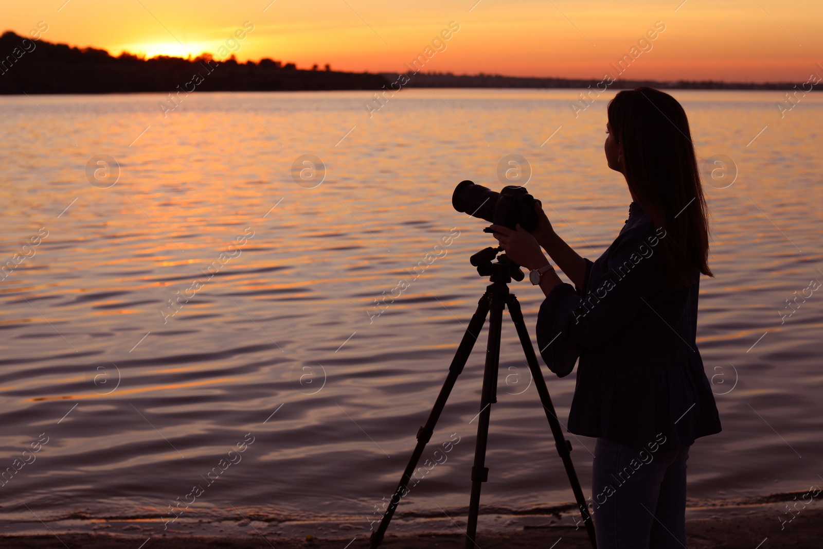 Photo of Young female photographer adjusting professional camera on tripod at sunset