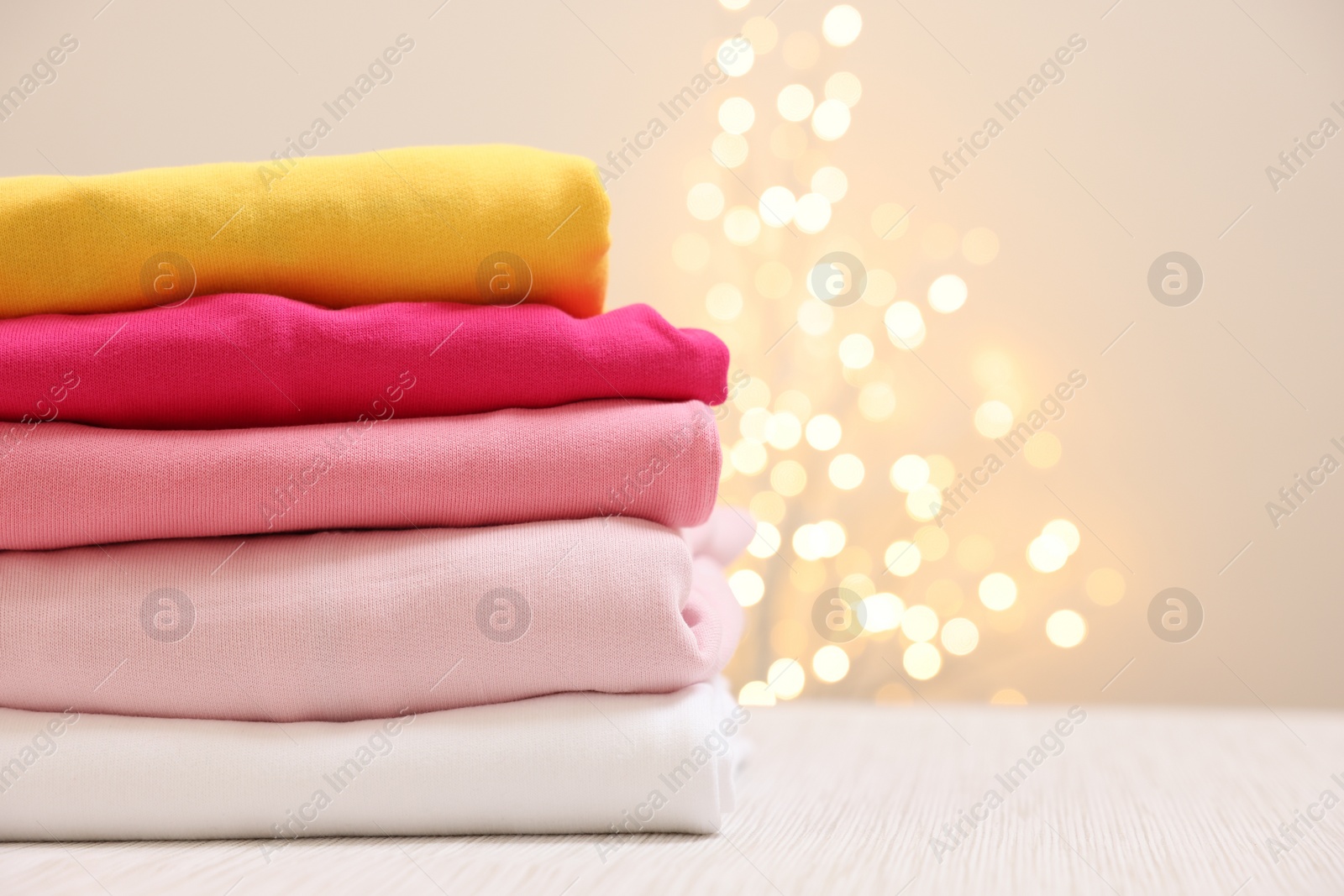 Stack of folded clothes on wooden table against beige background with blurred lights, space for text Photo of Stack of folded clothes on wooden table against beige background with blurred lights, space for text
