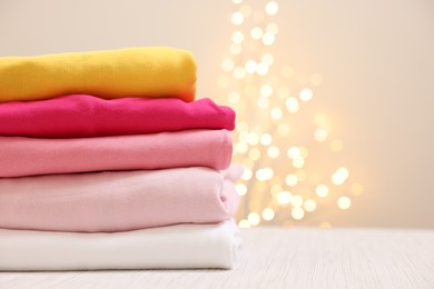 Photo of Stack of folded clothes on wooden table against beige background with blurred lights, space for text