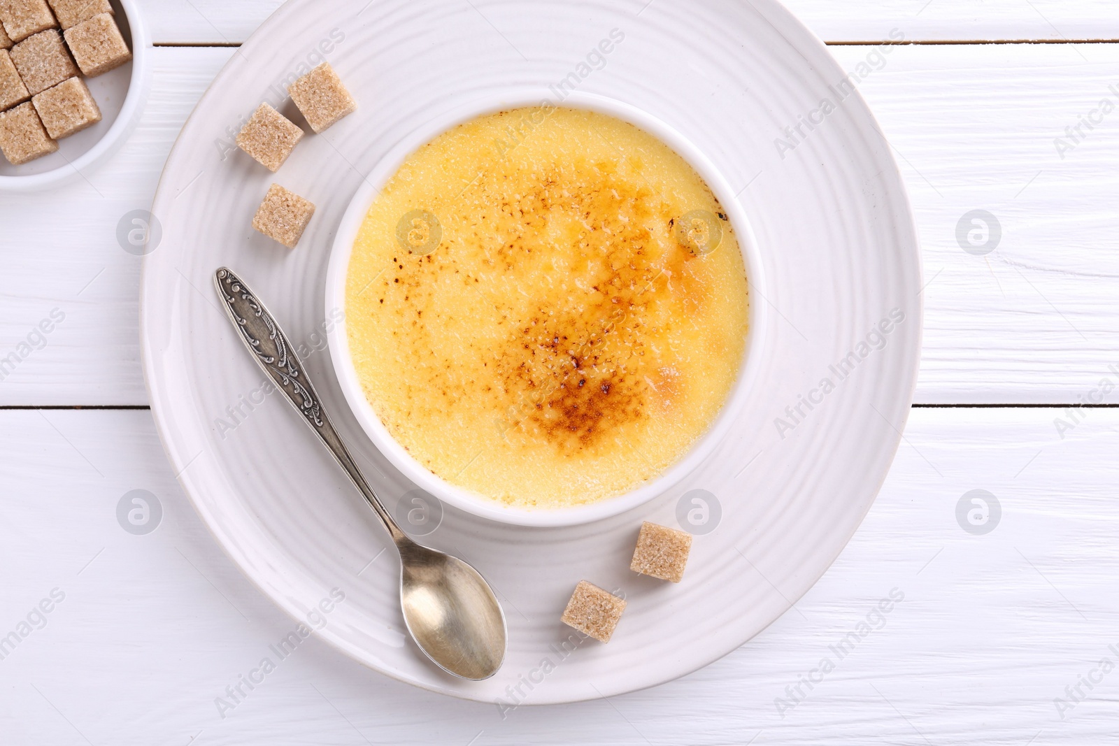 Photo of Delicious creme brulee in bowl, sugar cubes and spoon on white wooden table, top view