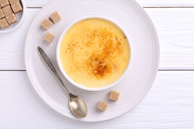 Delicious creme brulee in bowl, sugar cubes and spoon on white wooden table, top view