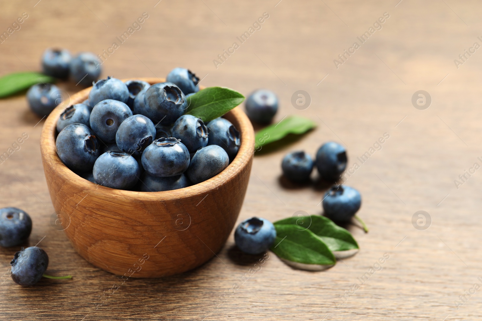 Photo of Bowl of tasty fresh blueberries with green leaves on wooden table, closeup. Space for text