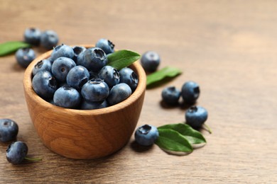 Bowl of tasty fresh blueberries with green leaves on wooden table, closeup. Space for text