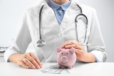 Doctor with piggybank and money at white table indoors, closeup. Medical insurance concept