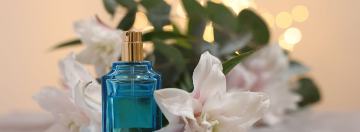 Bottle of perfume and beautiful lily flowers against beige background with blurred lights, closeup