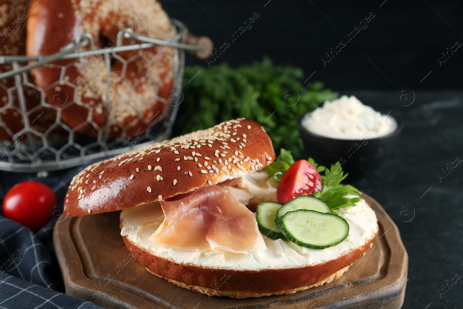 Photo of Delicious bagel with cream cheese, jamon, cucumber, tomato and parsley on black table