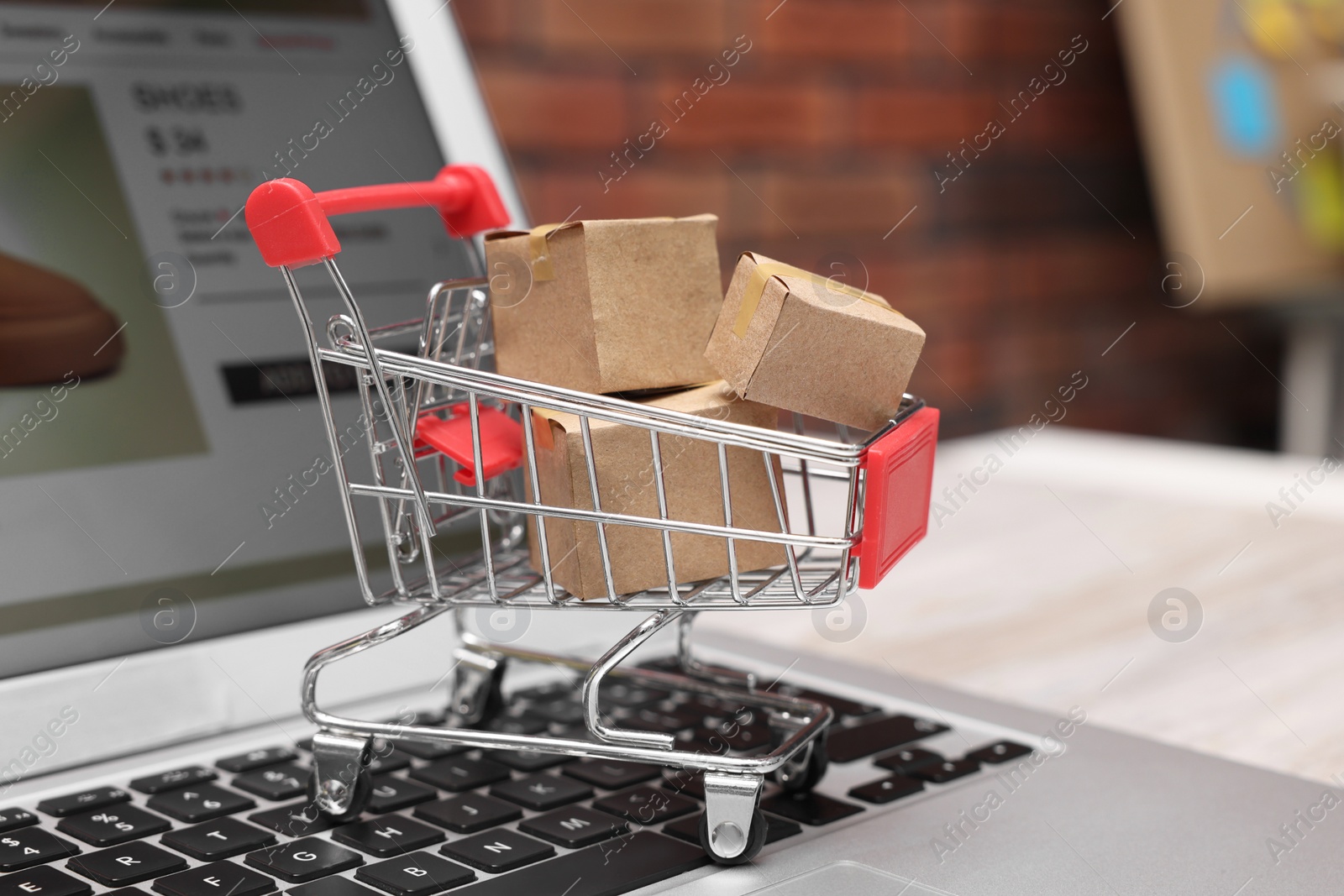 Photo of Internet store. Small cardboard boxes, shopping cart and laptop on light wooden table, closeup
