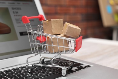 Internet store. Small cardboard boxes, shopping cart and laptop on light wooden table, closeup