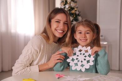 Happy mother and daughter with paper snowflake at table near Christmas tree indoors