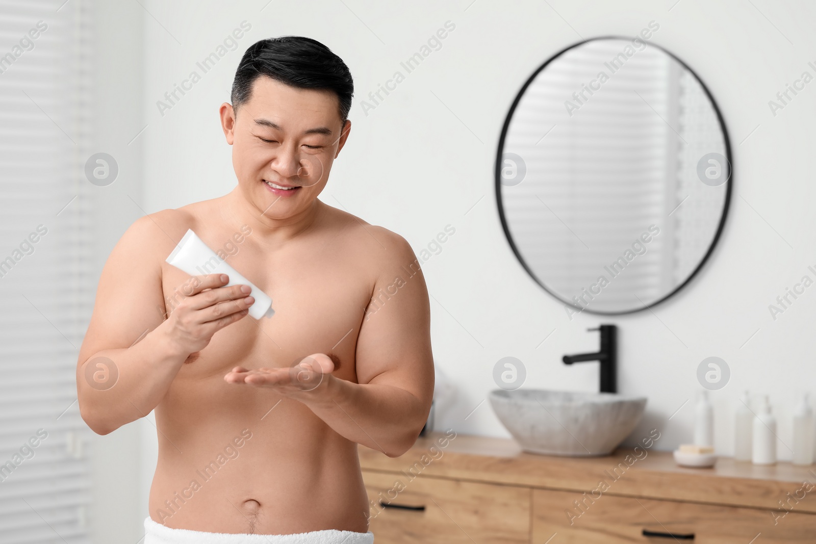 Photo of Handsome man applying body cream onto his hand in bathroom. Space for text