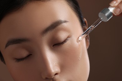 Photo of Beautiful young woman applying cosmetic serum onto her face on brown background, closeup