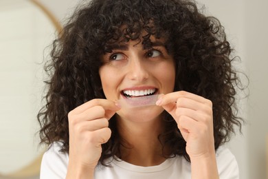 Young woman applying whitening strip on her teeth indoors
