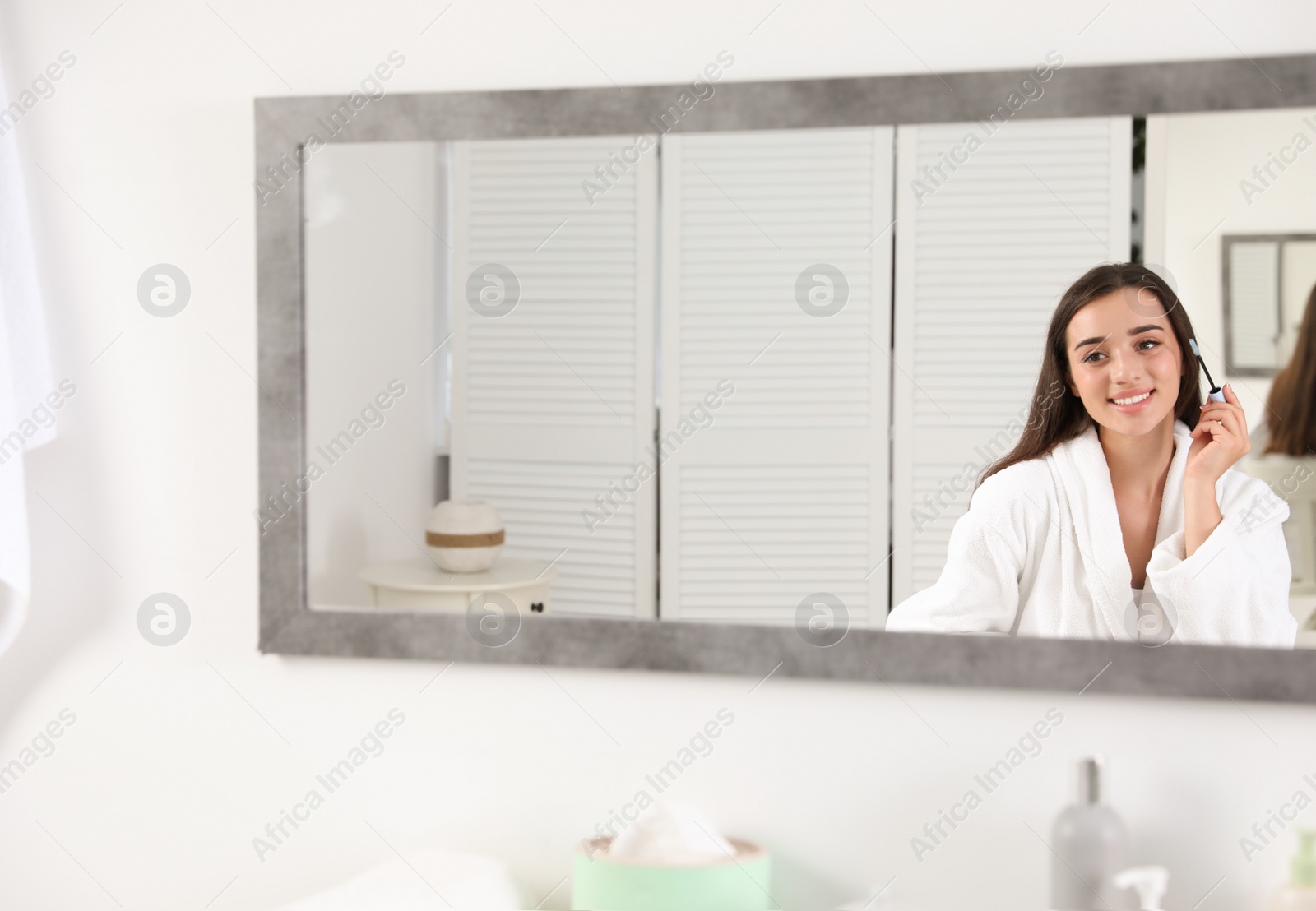 Photo of Attractive young woman applying oil onto her eyelashes near mirror indoors