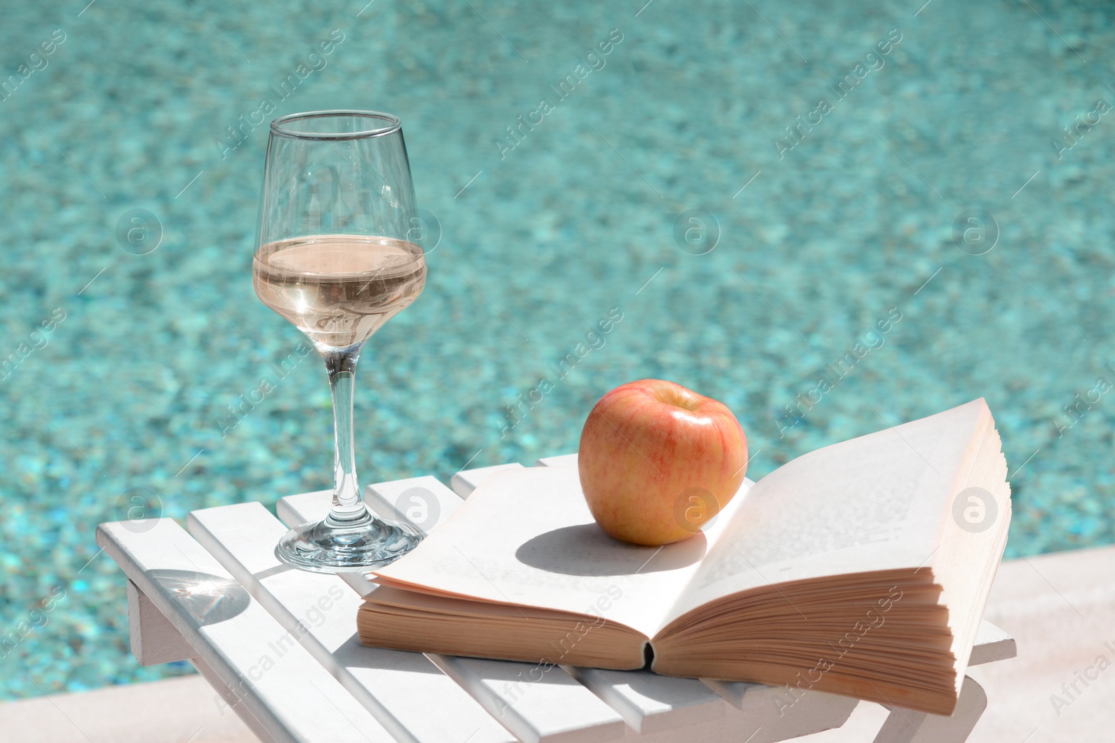 Photo of Glass of tasty wine, book and apple on wooden table near swimming pool