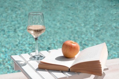 Photo of Glass of tasty wine, book and apple on wooden table near swimming pool