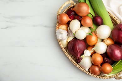 Photo of Wicker tray with fresh onion bulbs, leeks and garlic on white marble table, top view. Space for text