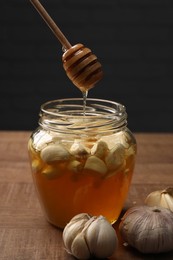 Photo of Natural honey dripping from dipper into glass jar with garlic on wooden table