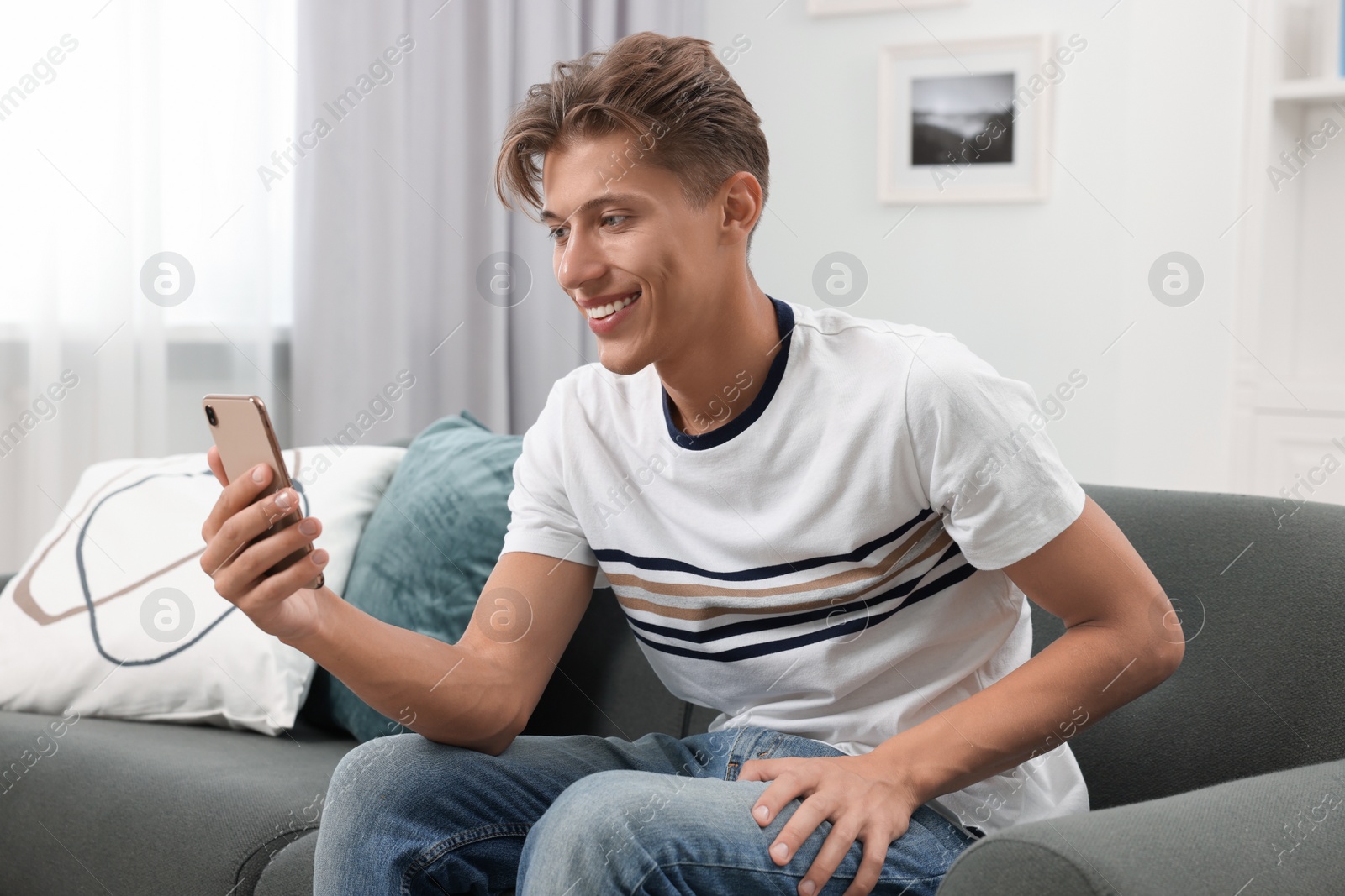 Photo of Happy young man having video chat via smartphone on sofa indoors