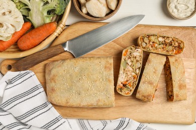 Photo of Cut tasty strudel with chicken, vegetables and knife on white wooden table, flat lay