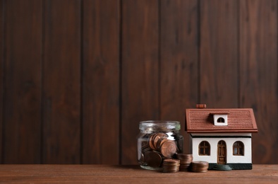 Photo of House model and jar with coins on table against wooden background. Space for text