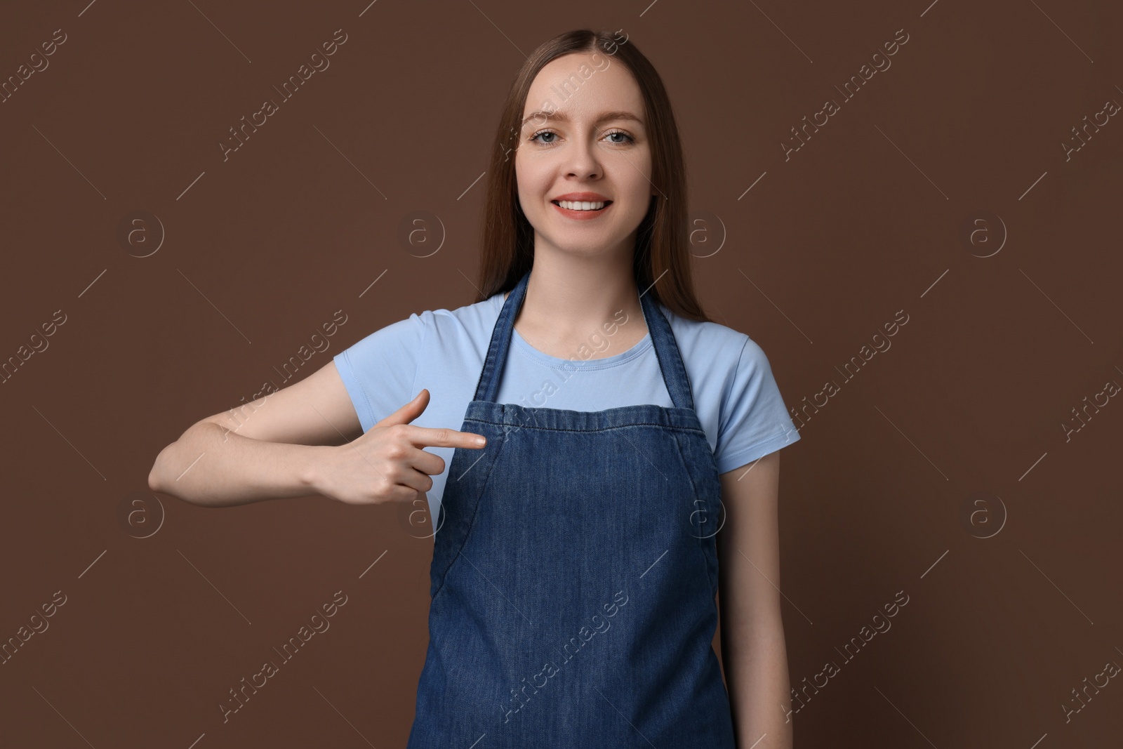 Photo of Beautiful young woman pointing at kitchen apron on brown background. Mockup for design