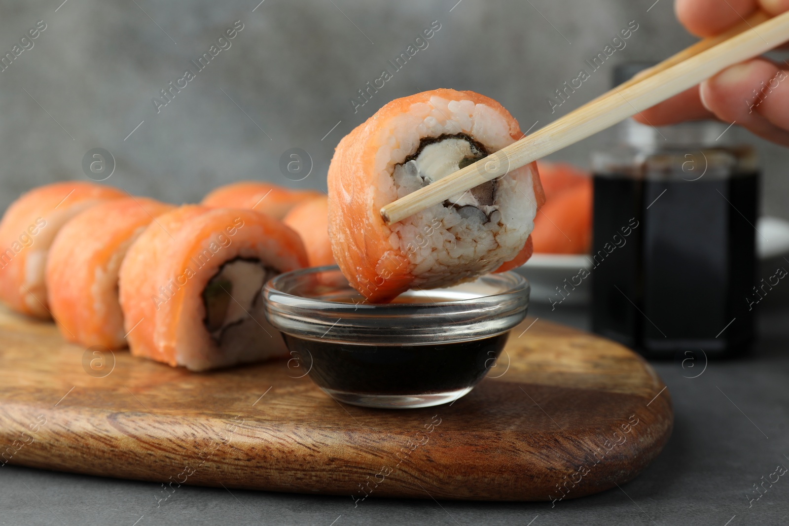 Photo of Woman dipping tasty sushi roll with salmon into soy sauce at grey table, closeup