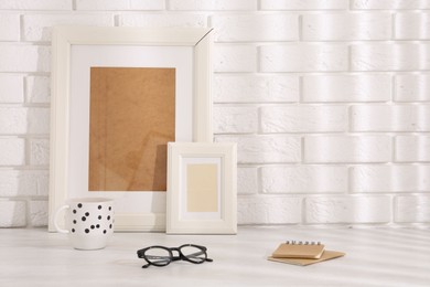 Stylish office workplace. Frames, glasses, cup and notebooks on white table near brick wall