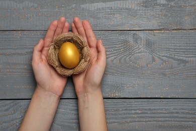 Photo of Woman holding nest with shiny golden egg at grey wooden table, top view. Space for text