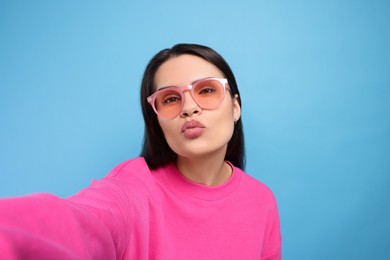 Beautiful young woman in stylish sunglasses taking selfie while giving kiss on light blue background