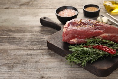 Photo of Piece of raw beef meat, rosemary, spices and chili pepper on wooden table, space for text