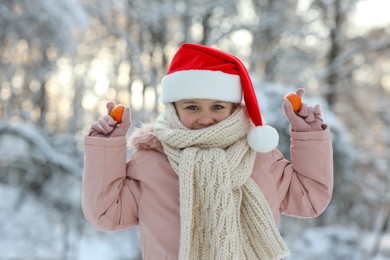 Cute little girl with tangerines wearing Santa hat in snowy park on winter day