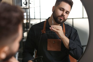 Photo of Handsome young man trimming beard with scissors near mirror indoors