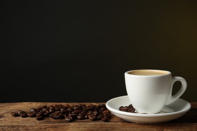 Photo of Cup of hot aromatic coffee and roasted beans on wooden table against dark background, space for text